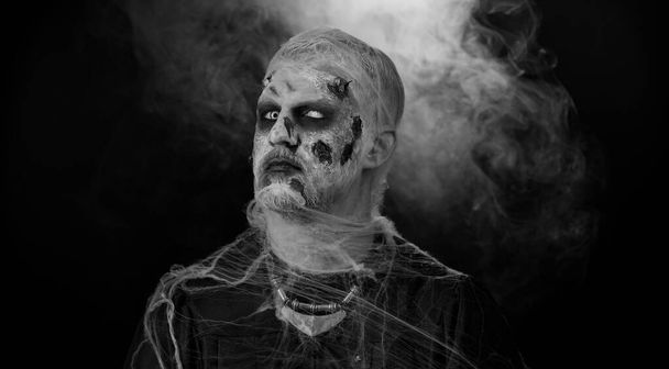 Cadılar Bayramı kostümlü uğursuz adam yaralı suratlı çılgın zombi suratlı bağıran, bağıran, siyah odayı korkutmaya çalışan. Kostümün korku teması yaralı ölümsüzler, canavar, canavar - Fotoğraf, Görsel