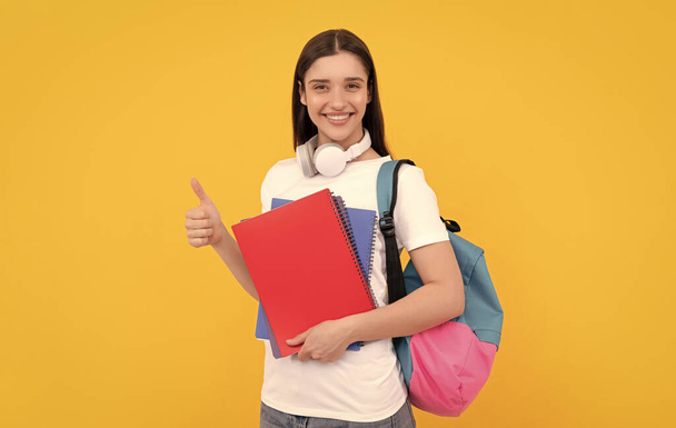 happy girl holding notebook on yellow background. private teacher with copybooks and headphones. back to school. modern education concept. thumb up. student hold notebook. - Photo, image
