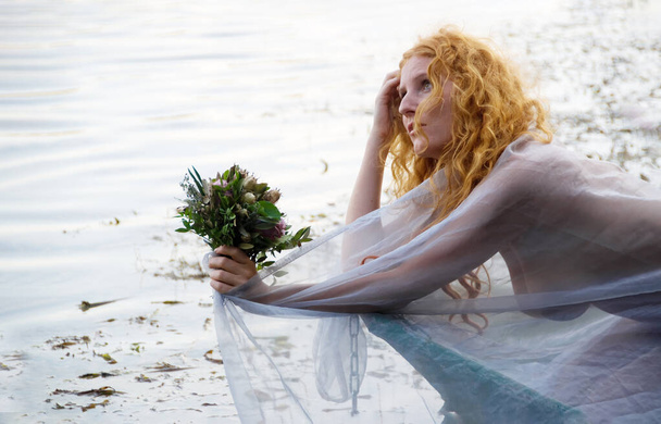 beautiful sensual, seductive, young sexy redhead woman with red hair, a longing, bride, Lady of Shalott, with a bouquet of wilted flowers lying in a boat in lake water, copy space - Фото, зображення