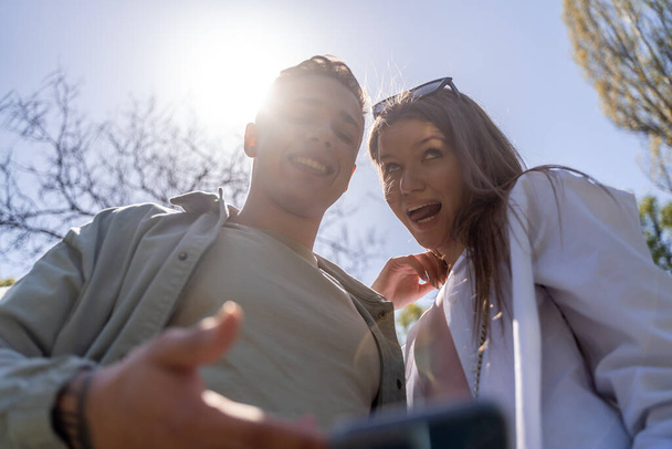 Dos amigos compartiendo contenido en línea en su teléfono inteligente. Pareja haciendo foto selfie en el parque - Foto, Imagen