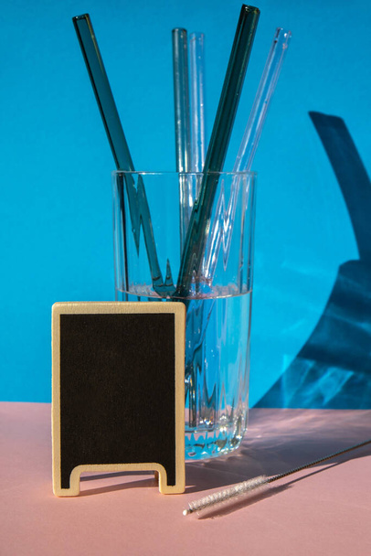 Pajitas de vidrio reutilizables en vidrio con agua sobre fondo colorido con etiqueta para su texto Set de pajitas ecológicas para beber con cepillo de limpieza. Cero residuos, concepto libre de plástico. Estilo de vida sostenible - Foto, imagen