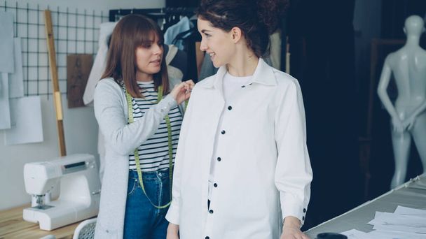 Pretty customer is trying on tailored shirt and sharing her opinion while female clothing designer is measuring and checking new garment and taking to client in light studio. - Photo, image