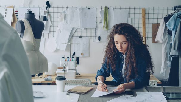 Young woman fashion designer is drawing ladies garment sketch at desk in modern workshop. Mannequin, sewing items, to-go coffee are visible. Creativity in clothing manufacturing concept. - Foto, afbeelding