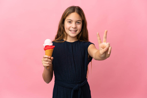 孤立したピンクの背景の笑顔と勝利のサインを示す上のコーネットアイスクリームを持つ子供 - 写真・画像