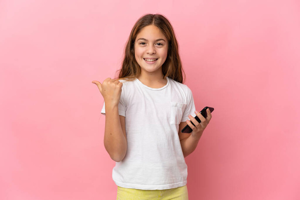 Child over isolated pink background using mobile phone and pointing to the lateral - Photo, image