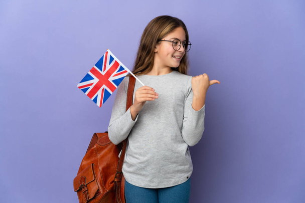 Kind hält eine britische Flagge über isoliertem Hintergrund und zeigt auf die Seite, um ein Produkt zu präsentieren - Foto, Bild