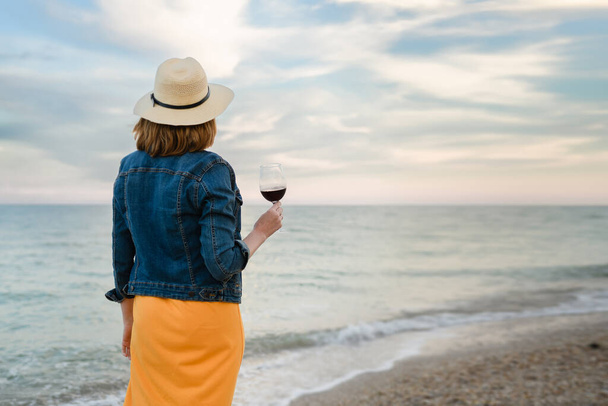 Young woman with a glass of red wine on the background of the sea. Evening sky. View from the back. Girl in a yellow dress and denim jacket. - 写真・画像