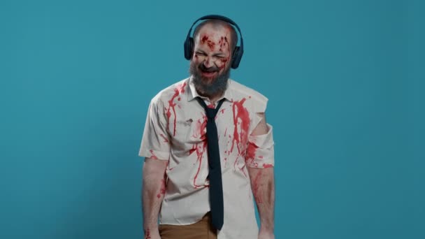 Грязный зомби носит электронные беспроводные наушники, слушая музыку на синем фоне. Зловещий апокалиптический монстр, поедающий мозг, наслаждающийся рок-радио, используя наушники. Снимок студии - Кадры, видео