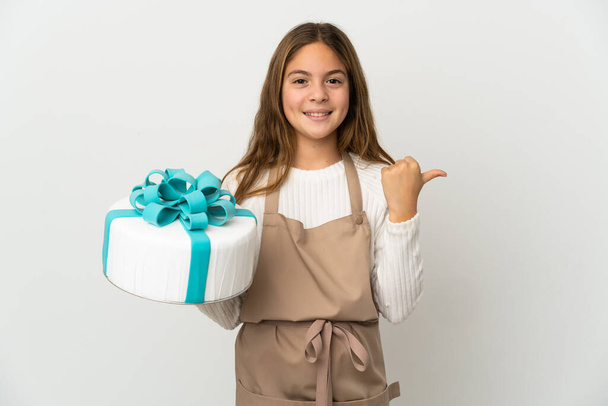 Klein meisje met een grote taart over geïsoleerde witte achtergrond wijzend naar de zijkant om een product te presenteren - Foto, afbeelding