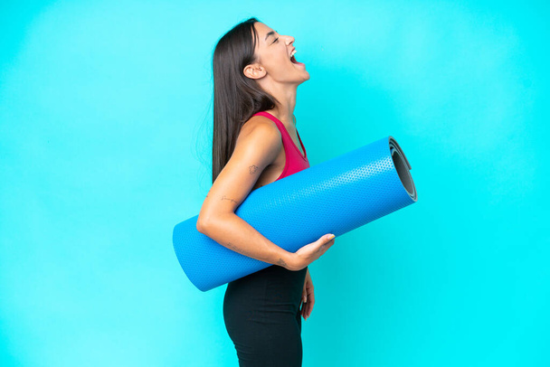 Junge Sportkaukasierin besucht Yoga-Kurse, während sie eine Matte isoliert auf blauem Hintergrund hält und in seitlicher Position lacht - Foto, Bild