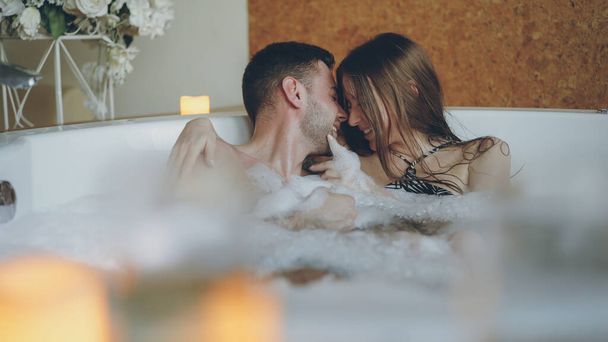 Adorable cheerful young couple is having fun in bubbling hot tub playing with foam, talking and kissing. Romantic relationships, happy people and wellness concept. - Fotografie, Obrázek