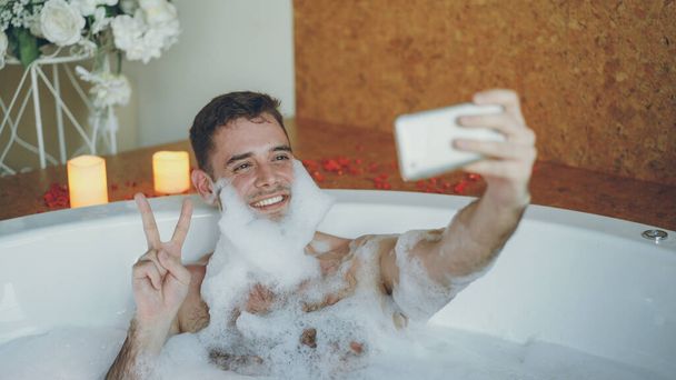 Guapo chico alegre se está tomando selfie en la bañera con espuma de jabón en la barba usando el teléfono inteligente. Se ríe y hace gestos con su mano posando y divirtiéndose
. - Foto, imagen