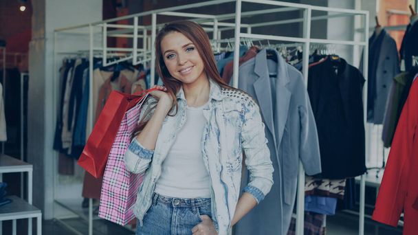 Portrait of happy and attractive girl standing in clothing shop with colourful bags, smiling gladly and looking at camera. Modern clothes is hanging in background. - Photo, Image