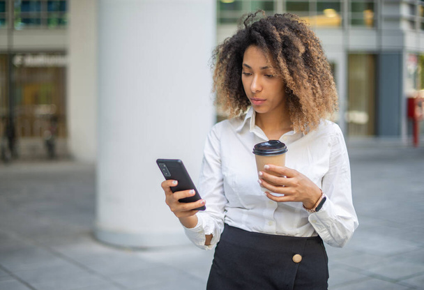 Portrait of a afro american business woman using her mobile phone in a business environment while drinking coffee - Foto, Imagem