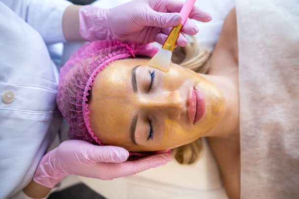 A beautician applies a golden mask made of medicinal oak bark to a woman's face. Cosmetology and facial skin care in a beauty salon. Cosmetic procedure. beautiful patient - Photo, Image