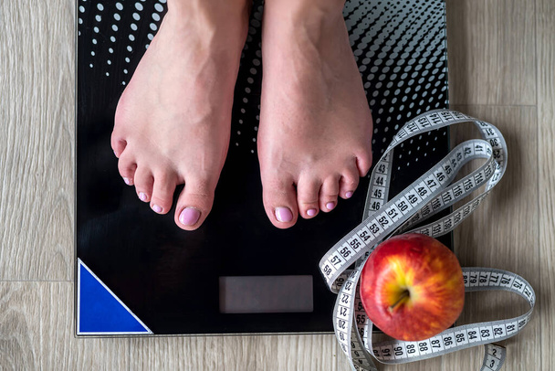Snímek ženy stojící na stupnici proti měřicí pásce a jablku. Nohy omotáme páskou. měření tělesné hmotnosti. samičí chodidla - Fotografie, Obrázek