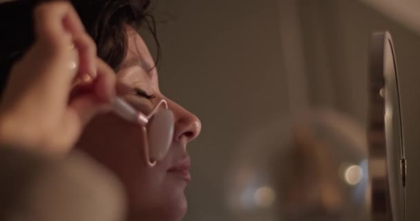 Low angle of brunette looking in round mirror and doing facial massage with jade roller during skin care routine in evening at home - Séquence, vidéo