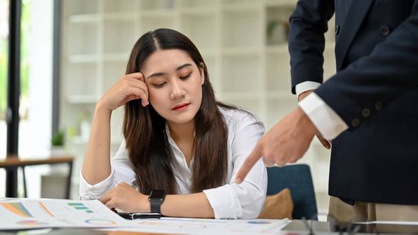 A férfi főnök panaszkodik egy fáradt és unatkozó fiatal ázsiai női könyvelő vagy női pénzügyi dolgozó az irodában. - Fotó, kép