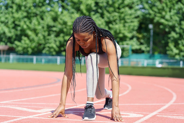 Black young sportive female athlete prepares low start running action on track of city stadium against green trees in park. African American woman with long braids trains before Olympics on sunny day - Fotografie, Obrázek
