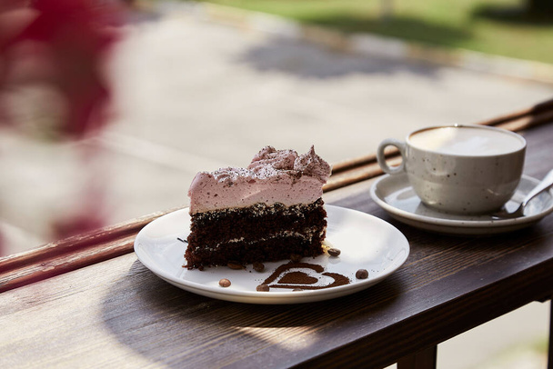 Atmosperic natural piece of cake and a glass of cappuccino on wooden table outdoor in the cafe terrace. Aesthetic coffee time. Time for yourself. Hard shadows. - Fotó, kép