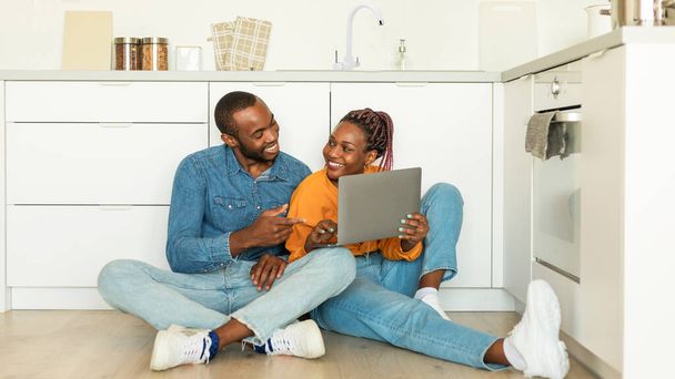 Happy black wife and husband using laptop, watch video or shopping online in kitchen interior, sitting together on floor. Video call, app and food blog - Fotografie, Obrázek