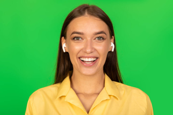 Headshot Of Joyful Lady Wearing Earbuds Earphones Listening To Music Smiling To Camera Posing Over Green Studio Background. Great Musical Application And Playlist Concept - Fotografie, Obrázek