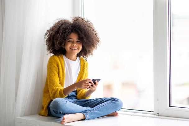 Cheerful cute african american little girl with bushy hair sitting on windowsill with smartphone, happy kid chatting with friends, using nice mobile application, home interior, copy space - Foto, Bild