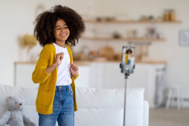 Pretty african american preteen girl with bushy hair in casual outfit recording video while dancing, showing dance moves, using cell phone on tripod, home interior, copy space. Kids lifestyle concept - Foto, Bild