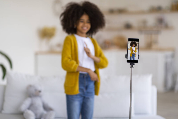 Selective focus on modern cell phone set on tripod recording video while cute curly preteen black girl in casual outfit dancing, having fun alone at home, blurred background, copy space - Foto, Bild