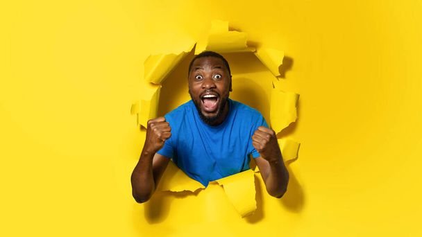 Overjoyed black man shouting happily, clenching both fists, cheering great success and victory, posing in ripped yellow paper hole, expresses positive emotions - Fotoğraf, Görsel