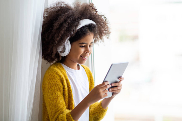 Closeup photo of cheerful black little girl sitting next to window at home, using brand new digital pad and white wireless headset, looking at tablet screen and smiling, watching cartoons, copy space - Foto, afbeelding