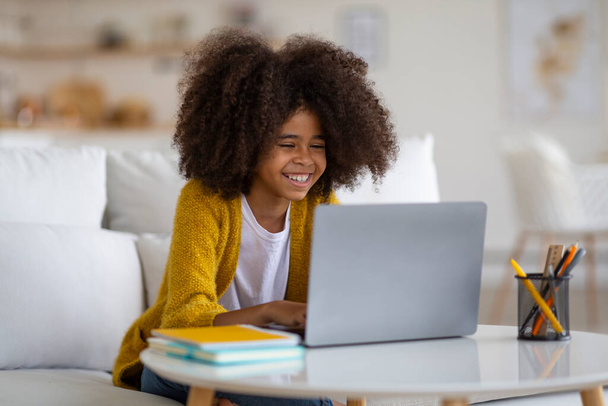 Smiling adorable black school girl doing homework, sitting on couch, using modern computer, typing on laptop keyboard, doing online test, home interior, copy space. E-education, homeschooling concept - Foto, immagini