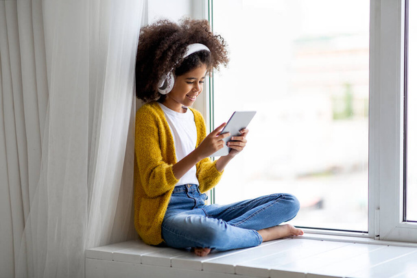 Cute african american school girl with bushy hair using modern digital tablet and wireless headset, sitting on windowsill at home, playing mobile game or checking newest app, copy space - Fotografie, Obrázek