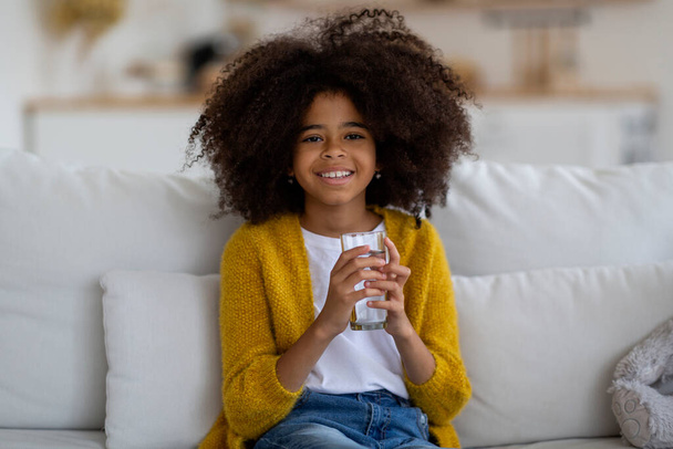 Cheerful pretty little black girl with bushy hair sitting on couch at home, holding glass of water and smiling at camera, kid drinking water, copy space. Hydration, children healthy lifestyle - Foto, immagini