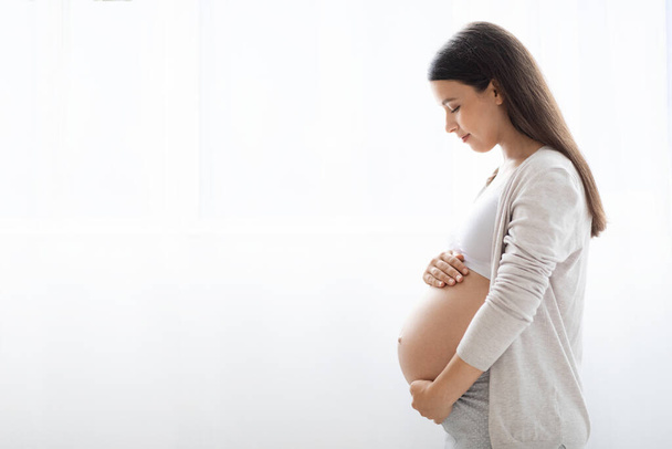 Side view of peaceful young expecting lady in comfortable homewear standing next to window at home, looking at her big tummy, waiting for baby, panorama with copy space. pregnancy concept - Foto, imagen