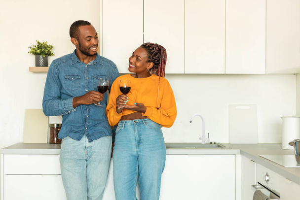Loving black couple drinking red wine at kitchen, happy man and woman standing with glasses, hugging and smiling at each other, free copy space - Фото, изображение