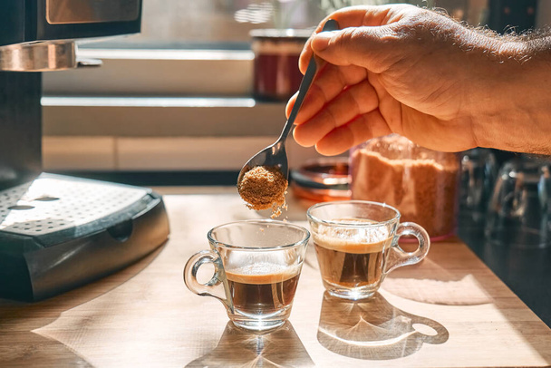 Kahverengi şekeri aromatik sıcak içecek espresso ile bardaklara koyan bir adam. Evdeki mutfakta espresso kahve makinesinde hazırlandı. Uyanış içkisi. Sabah alışkanlığı. - Fotoğraf, Görsel