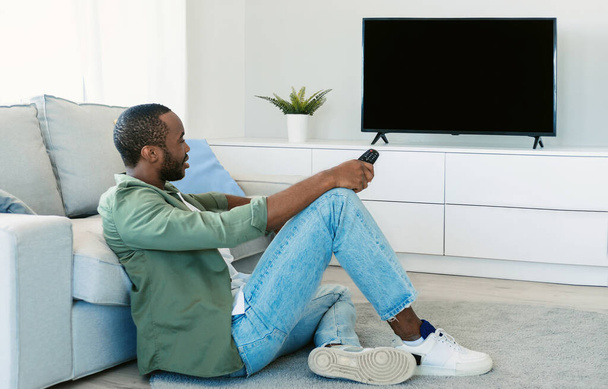 African american man watching television, turning on plasma flatscreen TV-set, pointing remote control at empty screen, mockup. Guy switching channels at home - Zdjęcie, obraz