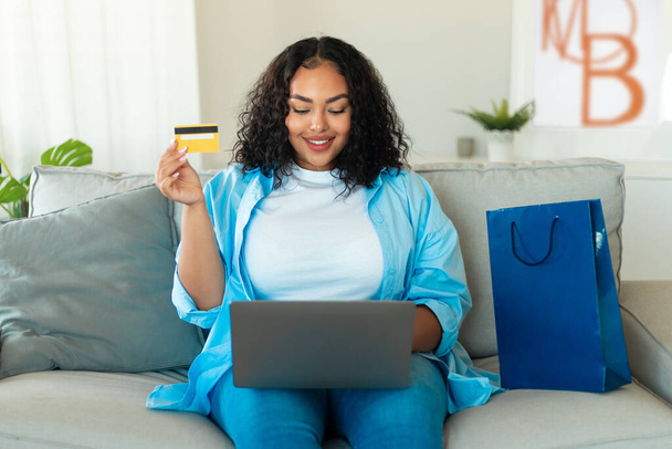 Електронна комерція. Африканська американська жінка використовує ноутбук і кредитну картку покупки онлайн на інтернет-ринку сидячи на дивані біля паперової сумки покупця вдома. Концепція веб банкінгу
 - Фото, зображення