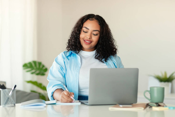 African American Lady Using Laptop And Taking Notes Working Online Sitting At Desk In Modern Office. Female Entrepreneurship And Business Career Concept. Selective Focus - Photo, image