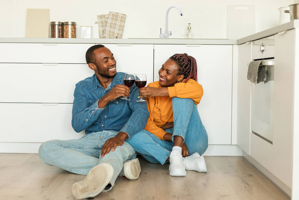 Romantic african american couple drinking red wine, sitting on floor in kitchen, enjoying time together and smiling to each other, celebrating Valentines day at home, copy space - Φωτογραφία, εικόνα