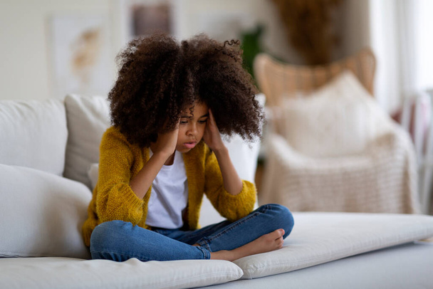 Unhappy little black girl with bushy hair suffering from headache, sitting on couch at home and touching her head, looking at copy space, wearing casual outfit. Kids health care concept - Foto, Imagem