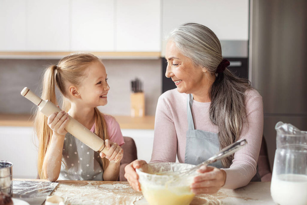 Cooking lesson. Smiling european little granddaughter and old grandma in aprons make dough with rolling pin, have fun in kitchen interior. Prepare homemade food together at home, love and relationship - Photo, Image