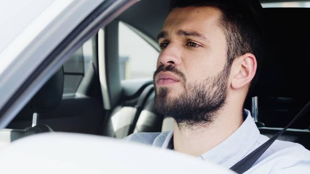 young and concentrated man looking ahead while driving car on blurred foreground - Foto, imagen