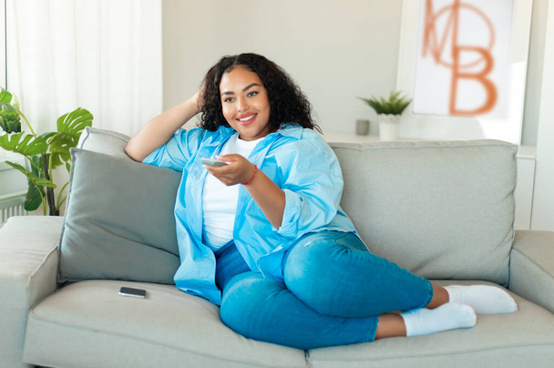 Cheerful Black Woman Turning On TV With Remote Controller Sitting On Sofa At Home. Female Viewer Watching Television On Weekend. Domestic Leisure And Entertainment - Photo, Image