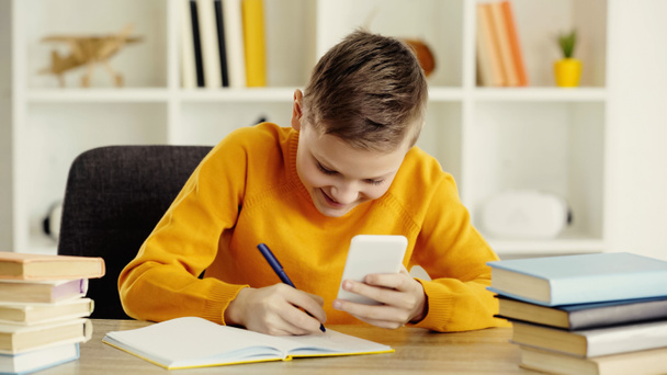 happy schoolboy using smartphone while writing in notebook near books on desk  - Fotó, kép
