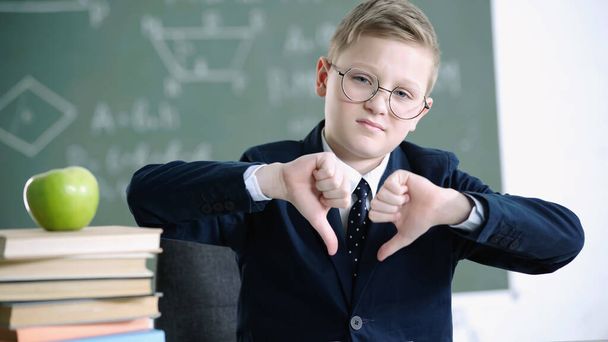 displeased schoolboy in glasses showing thumbs down near apple and books in school - Foto, Imagen