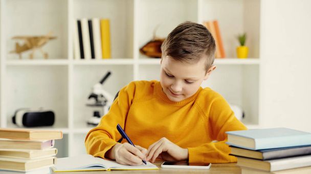 cheerful schoolboy writing in copy book near smartphone and books on desk  - Foto, Bild
