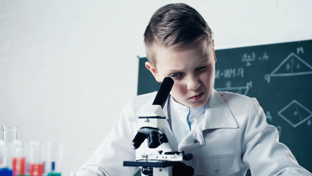 schoolboy in white coat looking through microscope during chemistry lesson in classroom   - Photo, image