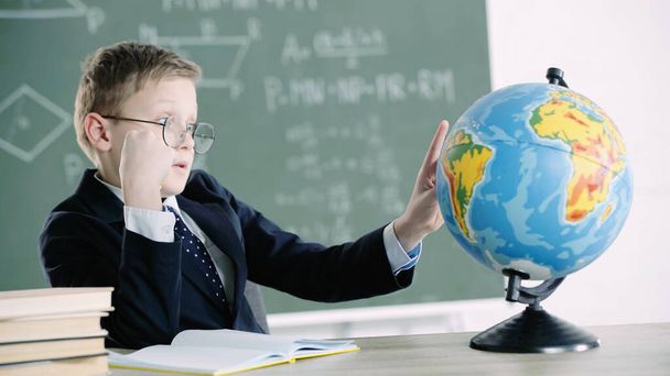 schoolboy in glasses pointing with finger at globe while sitting near books in classroom  - Photo, Image